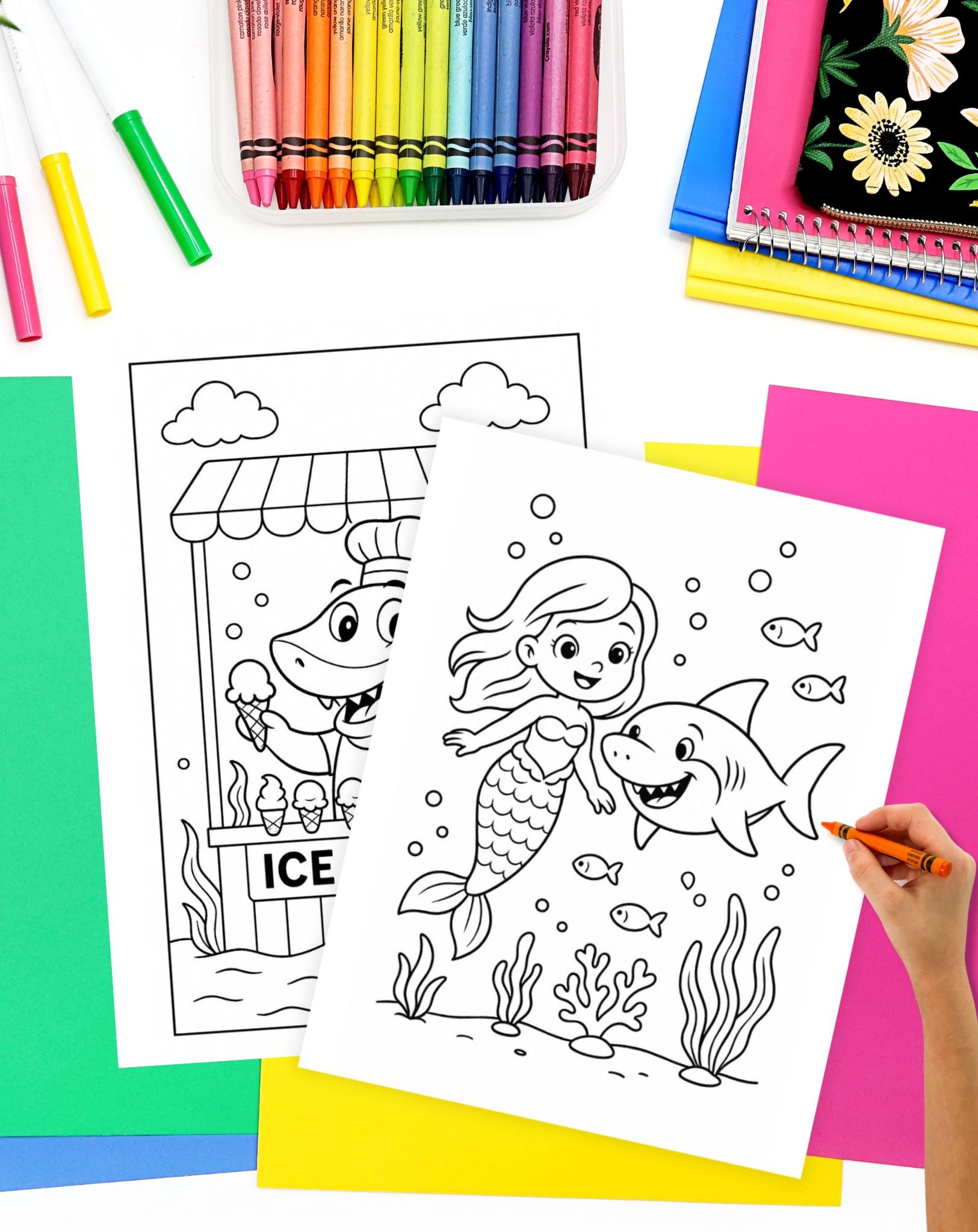 A hand is coloring a shark and mermaid illustration on a white sheet, surrounded by various ocean-themed coloring pages, crayons, and colorful paper sheets.
