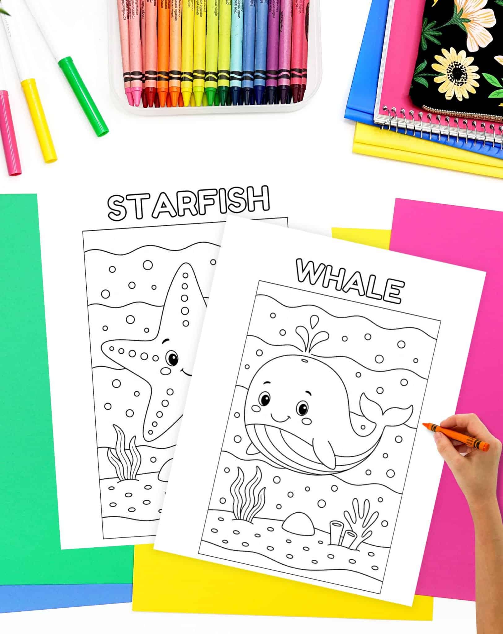 A hand is coloring a whale illustration on an ocean life coloring sheet, with a starfish coloring page also visible, surrounded by colorful crayons and various sheets of paper.
