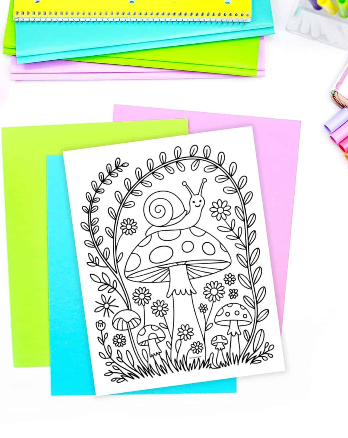 A black and white cottagecore coloring page featuring a snail, mushrooms, and flowers is placed on top of colorful sheets of paper, with additional stationery visible in the background.