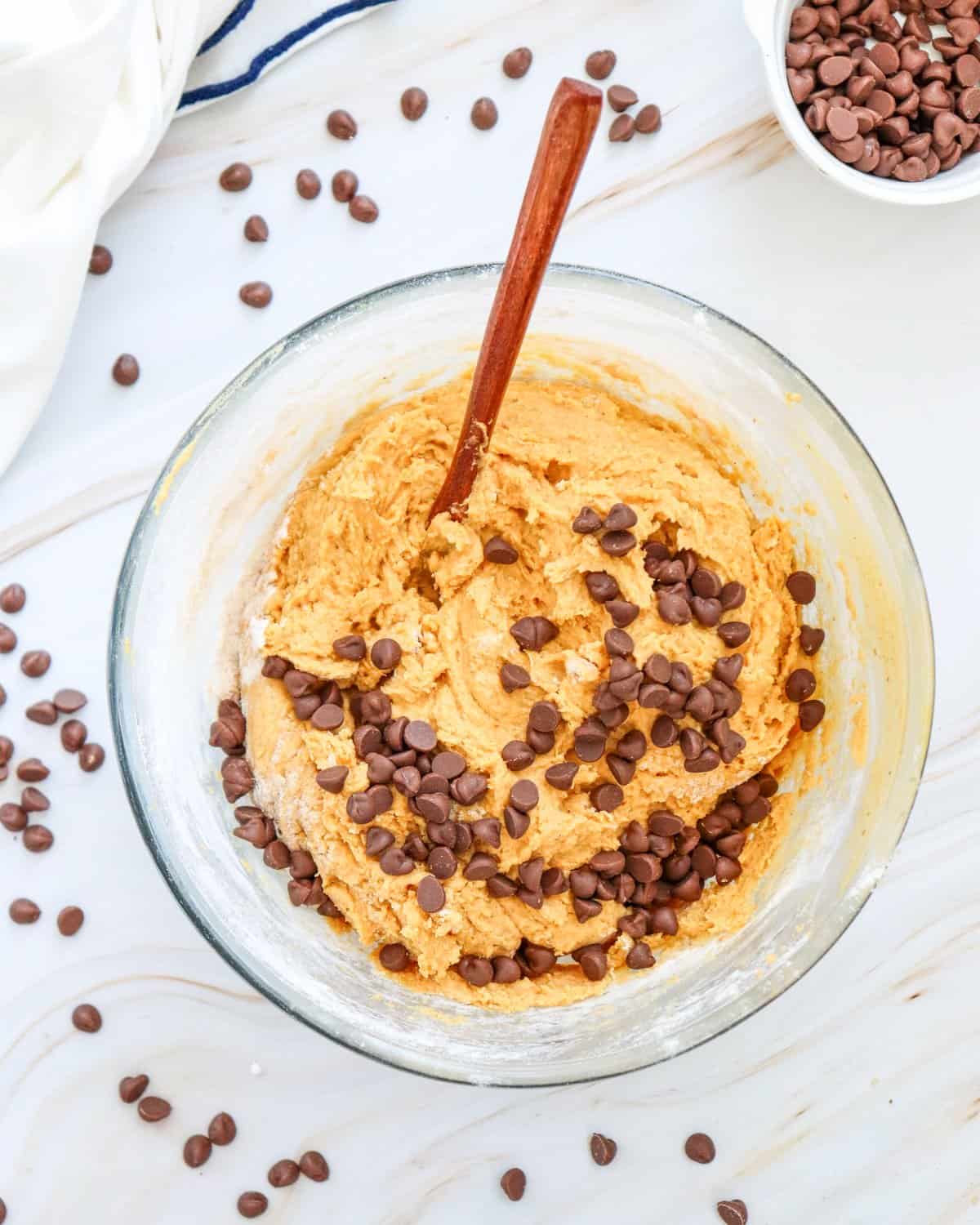 A glass bowl contains a mixture of cookie dough with pumpkin puree and an egg, alongside a hand mixer and a wooden spoon, surrounded by chocolate chips, illustrating the preparation for easy pumpkin cookies.
