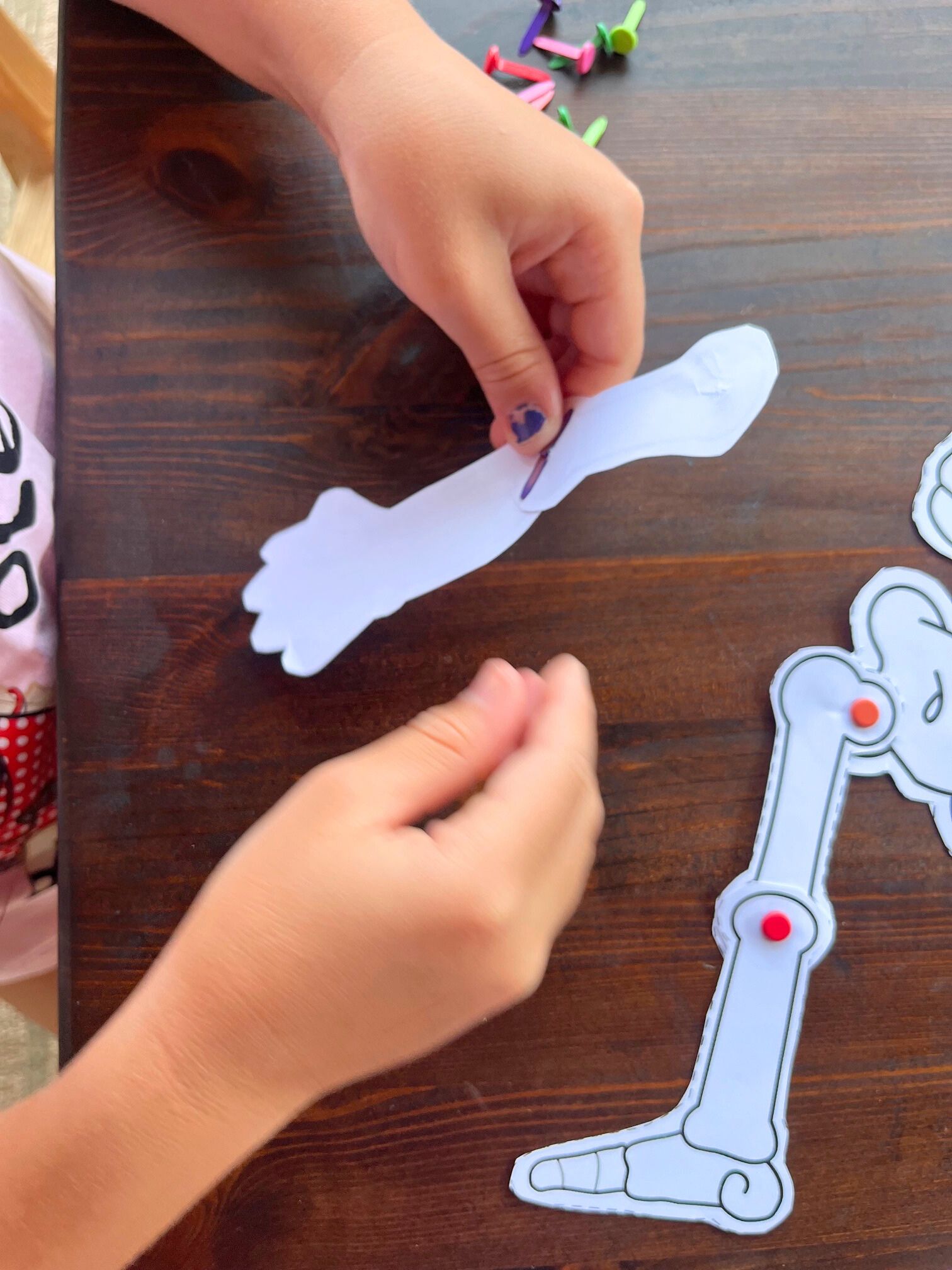 A child’s hands are assembling a printable skeleton template by attaching a paper arm to a pre-cut skeleton craft on a wooden table, surrounded by colorful push pins.
