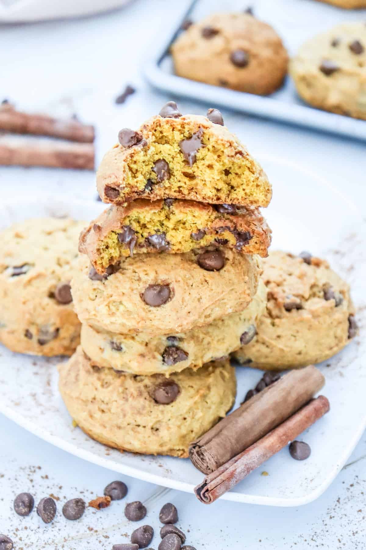 A stack of soft pumpkin cookies with chocolate chips is displayed, showcasing the texture and a few broken pieces on a plate with more cookies in the background.
