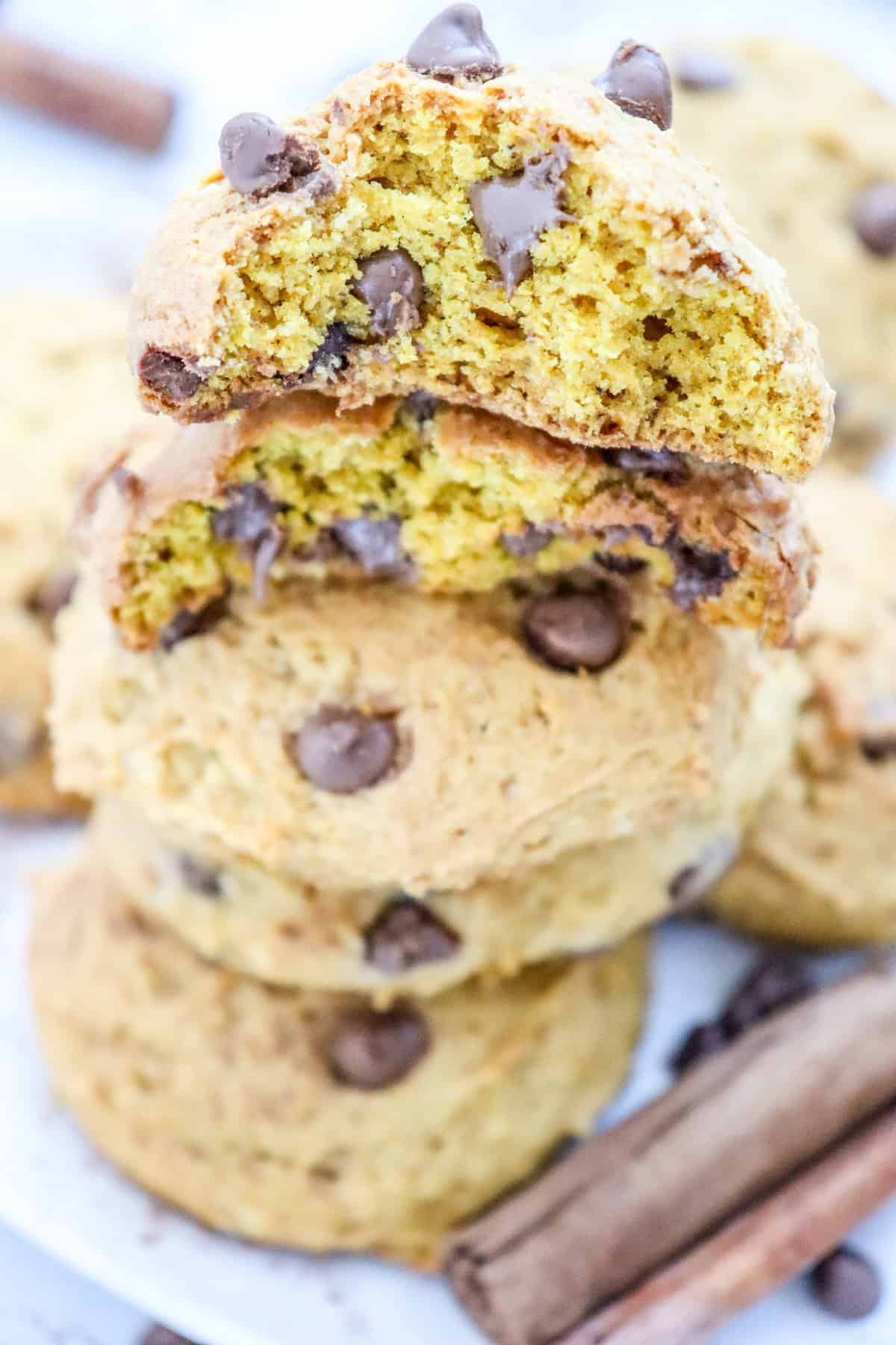 A stack of pumpkin cookies with chocolate chips is displayed on a plate, surrounded by cinnamon sticks and chocolate chips, highlighting a quick and easy pumpkin dessert recipe.

