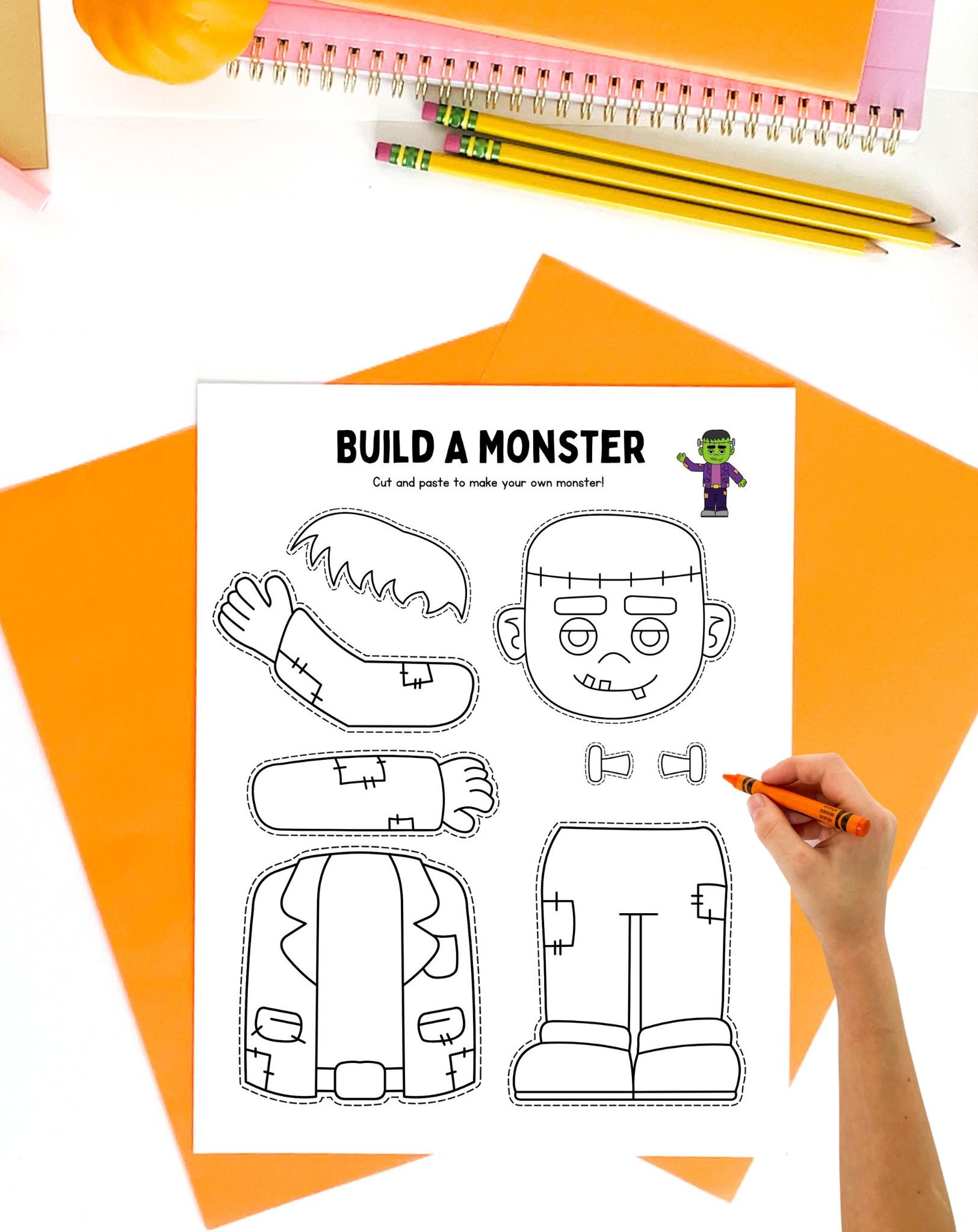 A child’s hand is coloring a "Build a Monster" printable, featuring cut-out parts for creating a Frankenstein craft, surrounded by colorful stationery on a table, ideal for Halloween classroom crafts and preschool activities.
