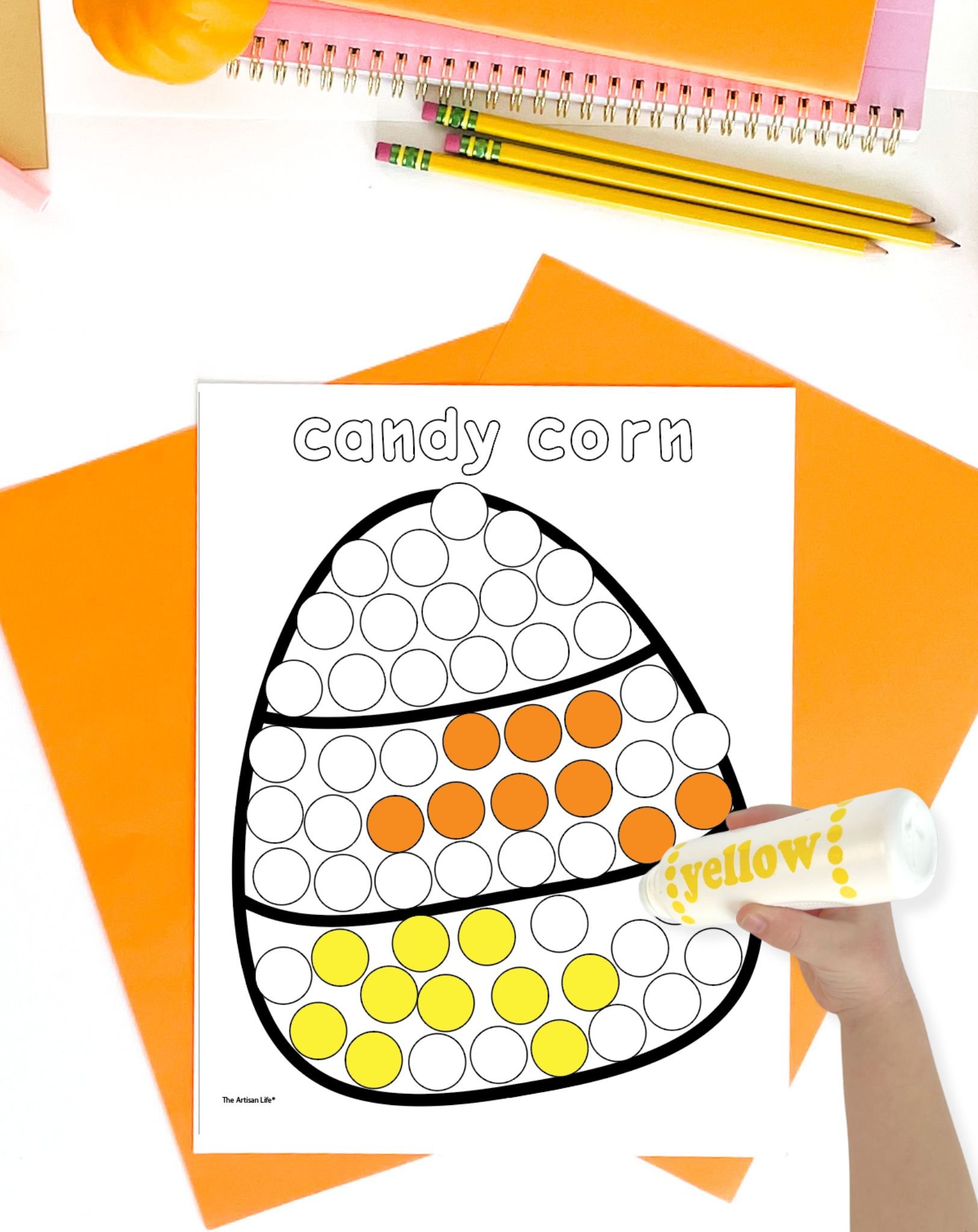 A hand is holding a yellow dot marker over a "candy corn" dot painting page on a table with orange and pink stationery, showcasing Halloween dot marker pages for Halloween activities.
