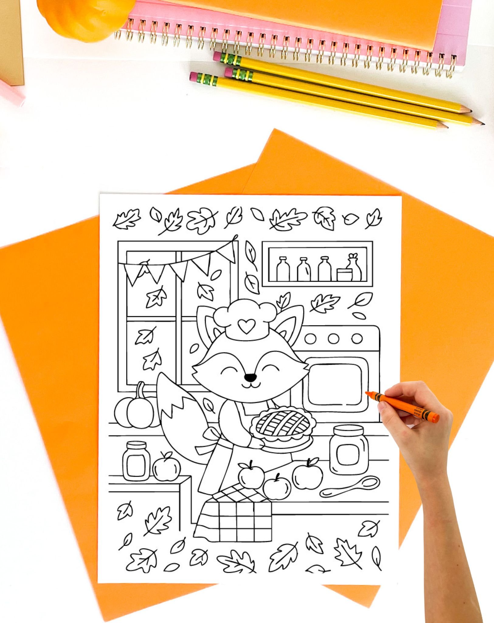 A hand holding an orange crayon colors a printable fall-themed illustration featuring a cheerful fox in a kitchen holding a pie, surrounded by autumn leaves, apples, and pumpkins, amidst a backdrop of cozy decor, representing free printable fall coloring pages.
