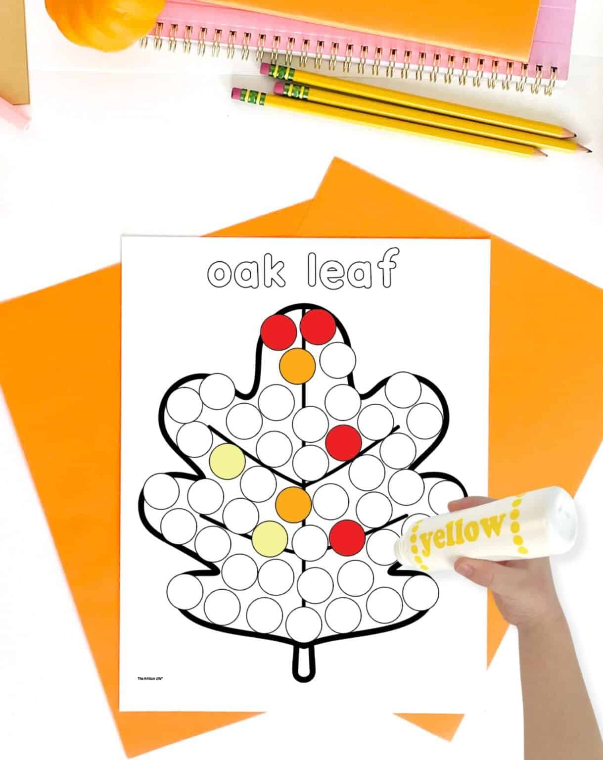 A hand is holding a yellow dot marker over a printable worksheet featuring an outlined oak leaf filled with numbered circles, on a backdrop of orange construction paper and stationery, illustrating a fall-themed dot marker activity suitable for preschool.