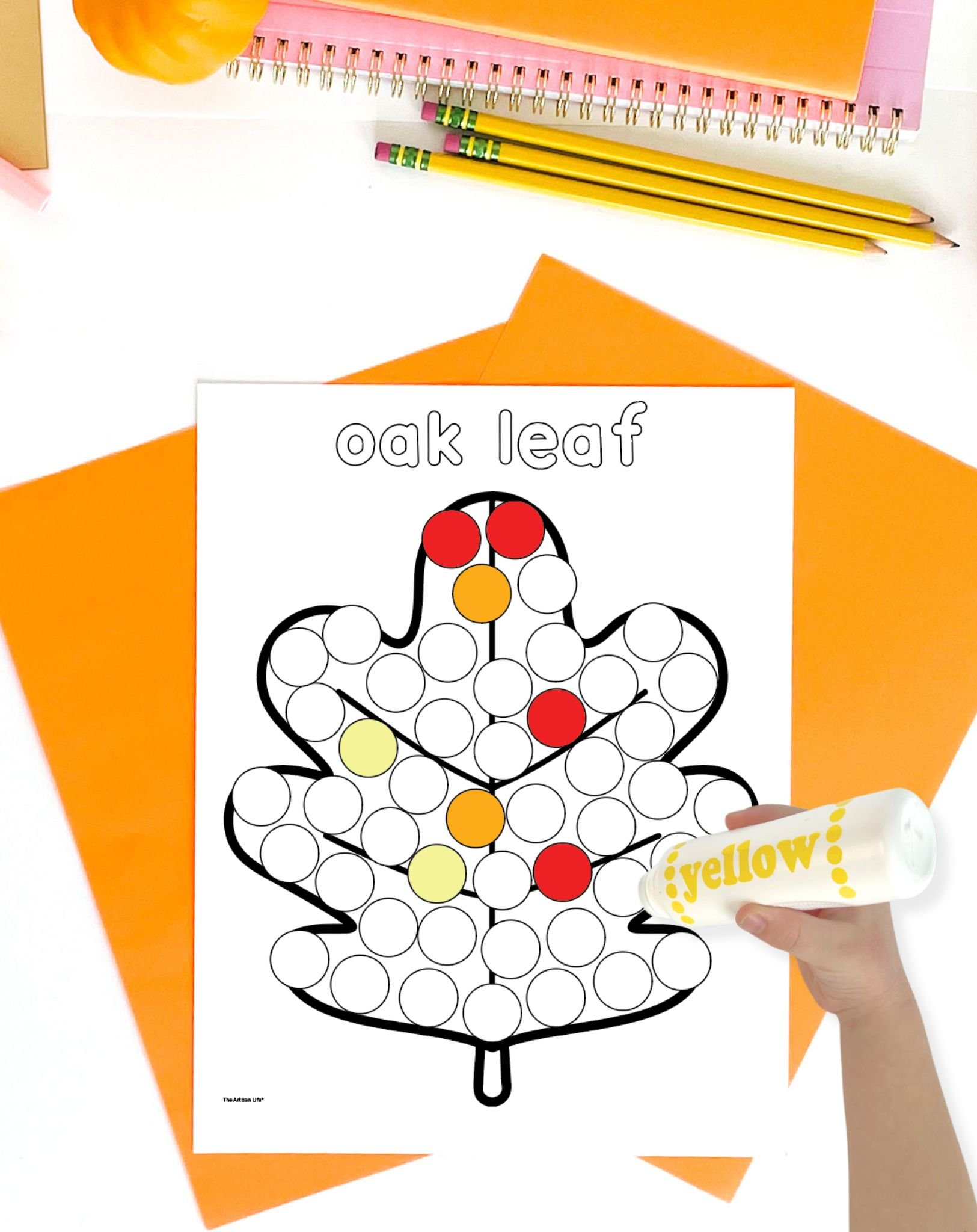 A hand is holding a yellow dot marker over a printable worksheet featuring an outlined oak leaf filled with numbered circles, on a backdrop of orange construction paper and stationery, illustrating a fall-themed dot marker activity suitable for preschool.
