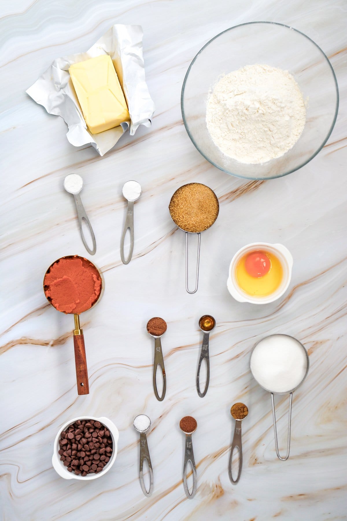 An assortment of ingredients for making pumpkin chocolate chip cookies, including butter, flour, pumpkin puree, chocolate chips, sugar, an egg, and various spices, is arranged on a marble surface.
