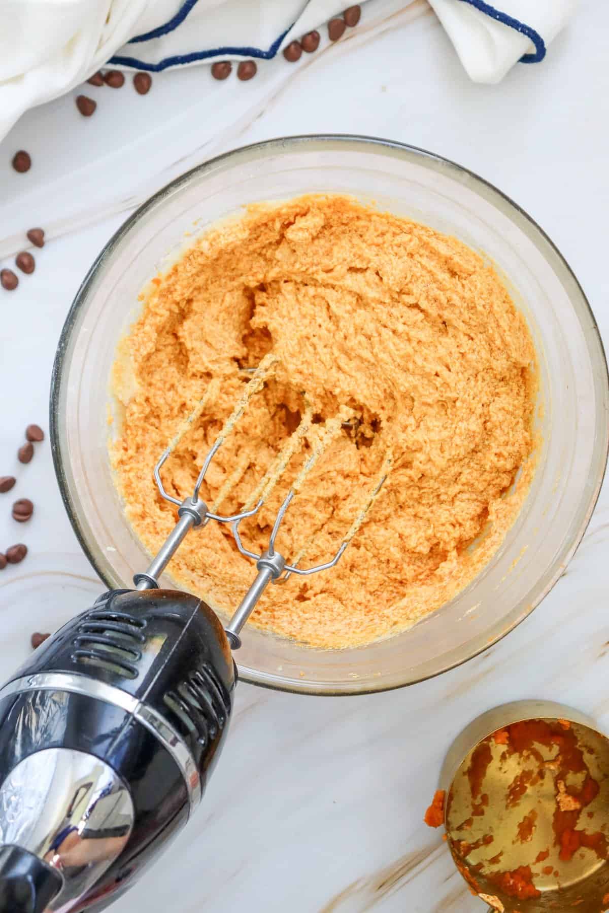 A glass bowl containing orange cookie dough is seen with an electric mixer resting beside it, accompanied by an open can of pumpkin puree and scattered coffee beans on a marble surface, suggesting preparation for pumpkin chocolate chip cookies or an easy pumpkin dessert recipe.
