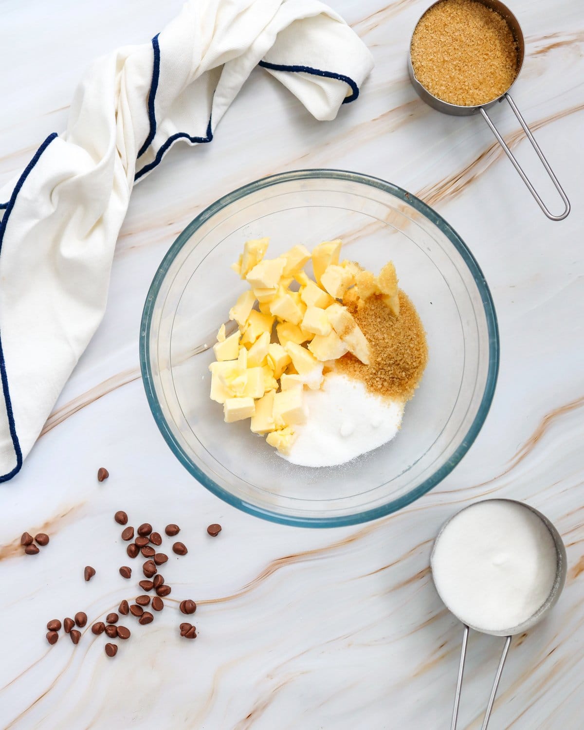A glass bowl contains chopped butter and various types of sugar, with scattered chocolate chips and measuring cups nearby, suggesting preparation for a pumpkin cookie recipe.
