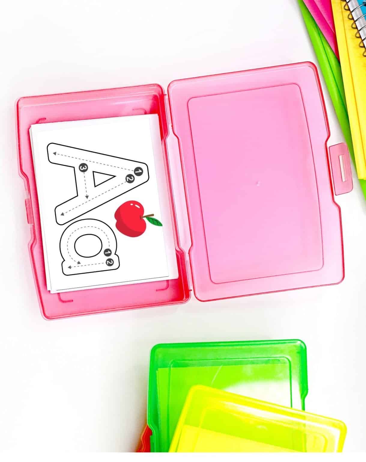 A pink storage case contains alphabet tracing cards featuring the letters "A" and "O" with accompanying images for letter formation practice, suitable for preschool handwriting activities.