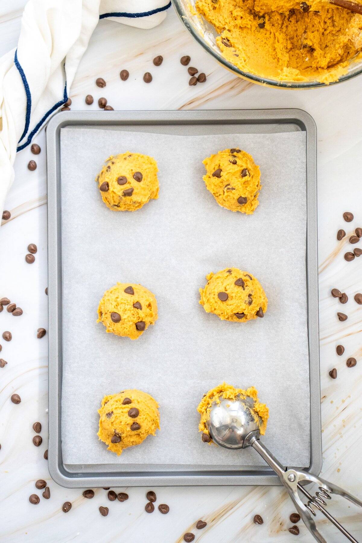 A baking sheet lined with parchment paper holds six dollops of pumpkin chocolate chip cookie dough, surrounded by scattered chocolate chips.
