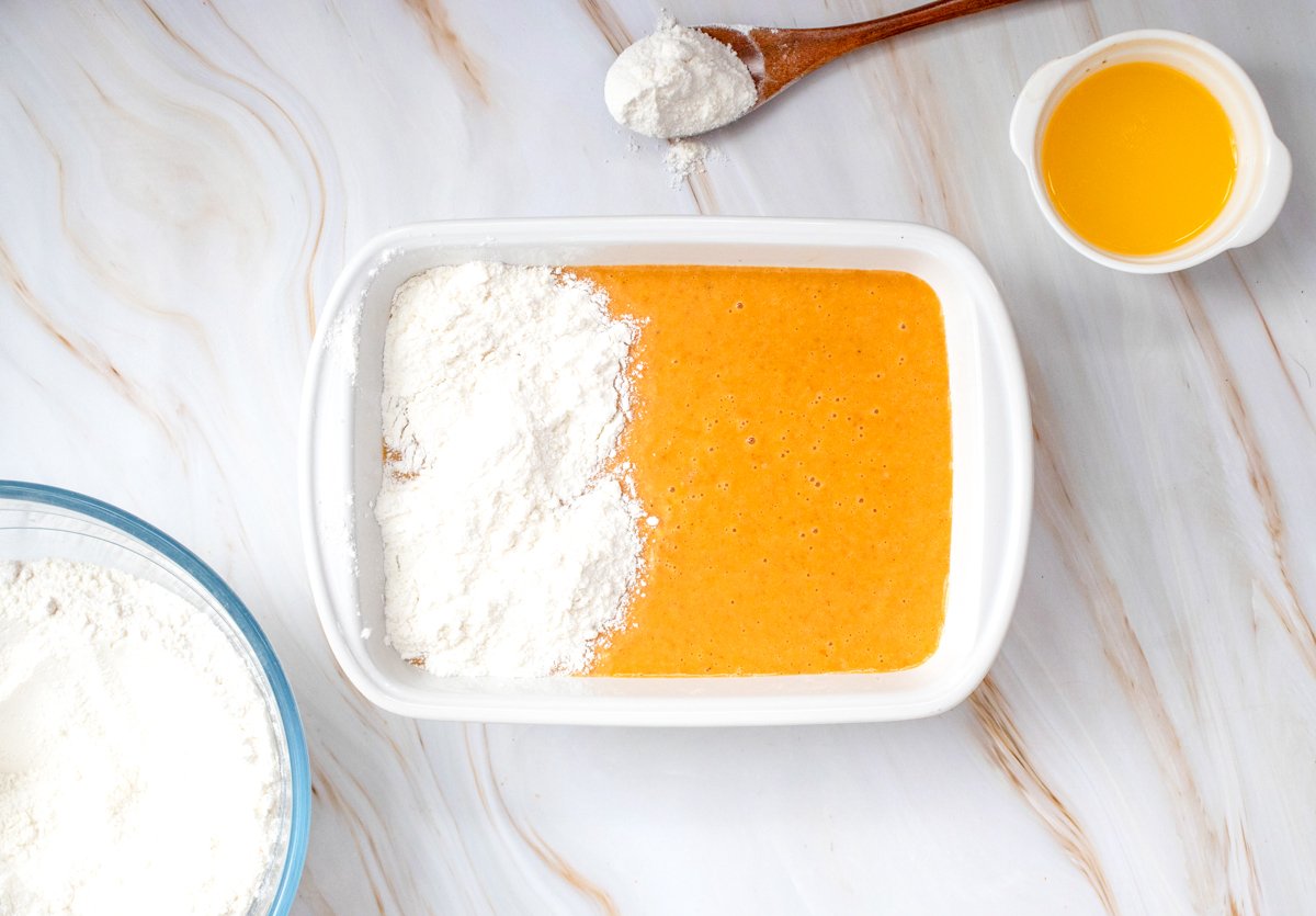 A baking dish with a mixture of pumpkin puree and eggs on one side and a pile of flour on the other, accompanied by a spoon of flour and a small cup of liquid, illustrates the preparation for an easy pumpkin dump cake recipe.
