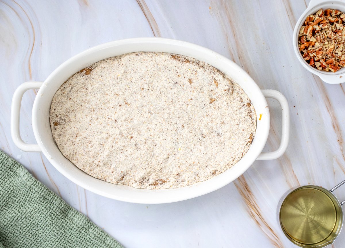 A white baking dish holds a mixture of pumpkin dump cake, surrounded by ingredients including gluten-free flour, chopped pecans, and a green kitchen towel on a marble surface.

