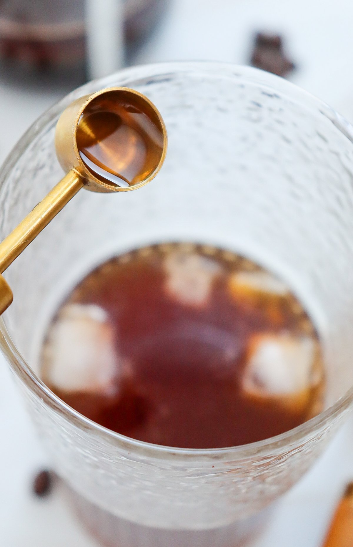 A close-up view of a glass filled with iced coffee, featuring a golden measuring spoon above it with caramel syrup, suggesting a recipe for homemade caramel iced coffee.
