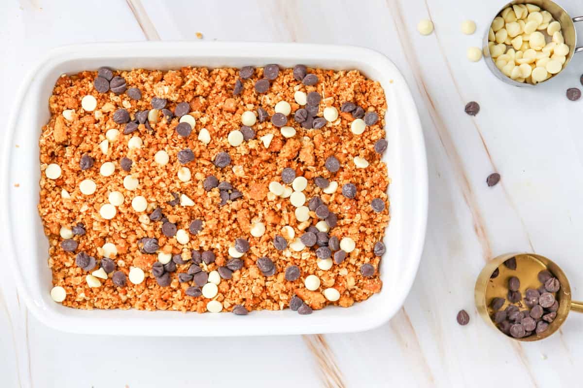 A white baking dish contains a layered pumpkin crisp topped with chocolate and white chocolate chips, surrounded by measuring cups of chocolate chips on a marble surface.
