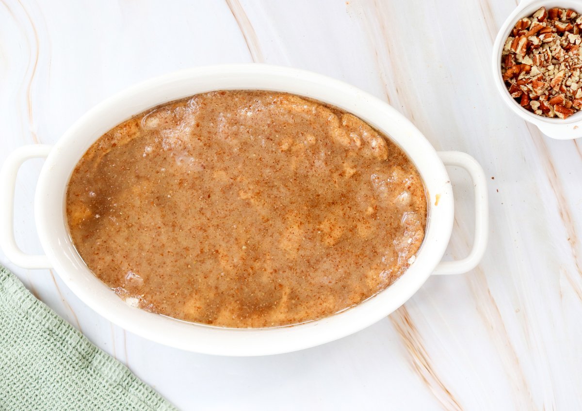 A white baking dish filled with uncooked pumpkin dump cake mixture sits on a marble countertop, with a small bowl of chopped pecans beside it, representing a gluten free dessert idea.
