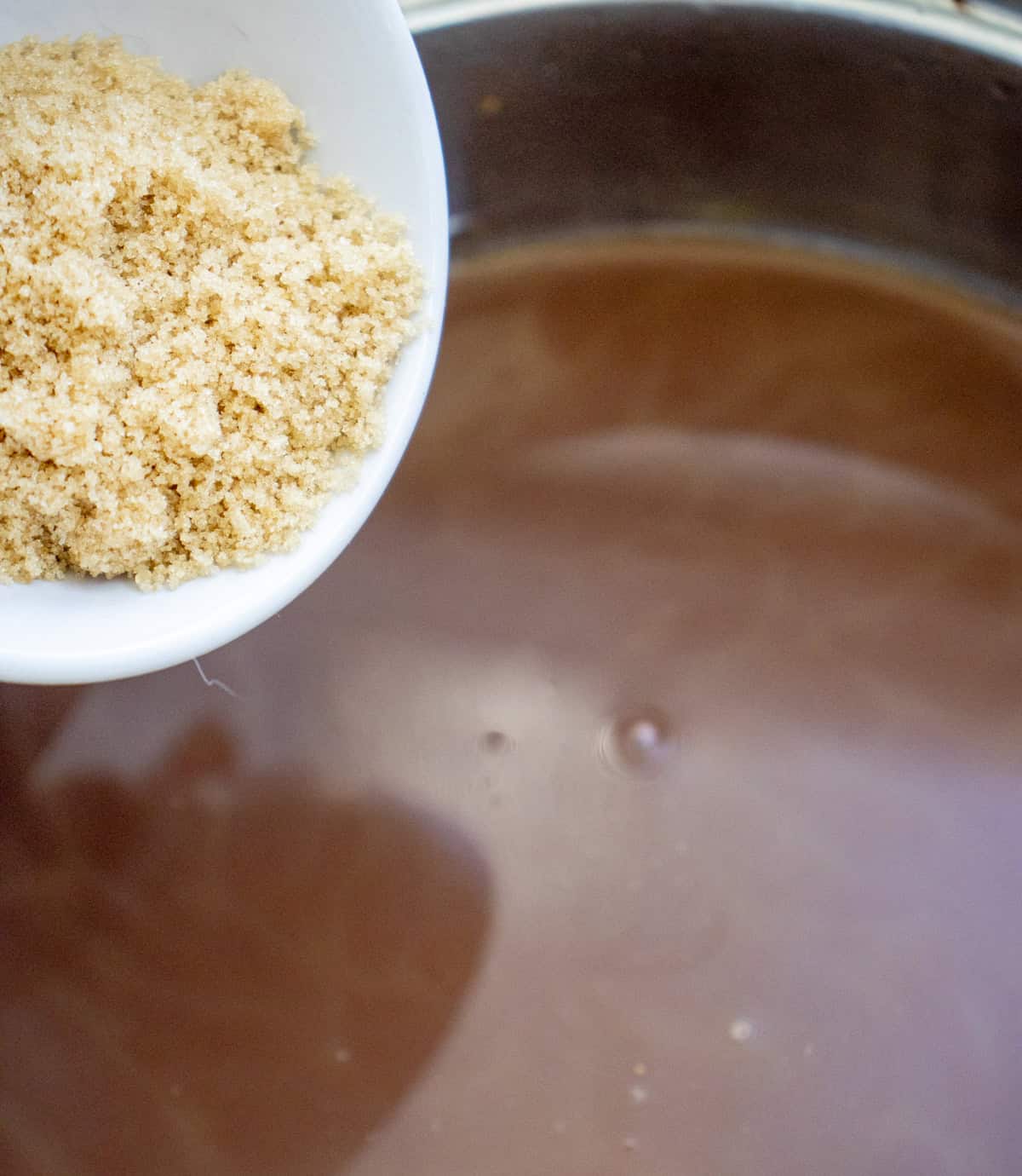 A hand holds a bowl of brown sugar above a pot of hot chocolate, suggesting preparation for fall drinks like pumpkin spice hot chocolate.
