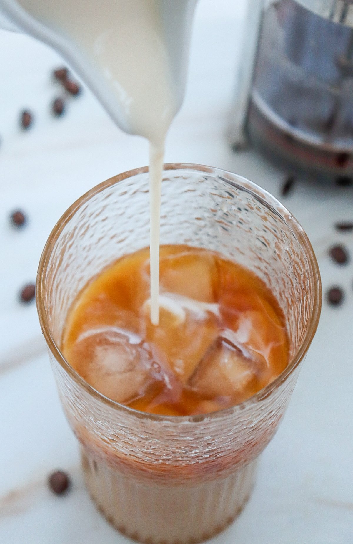 A glass of iced coffee with ice cubes is shown as cream is being poured in, surrounded by coffee beans, illustrating a homemade caramel iced coffee recipe.
