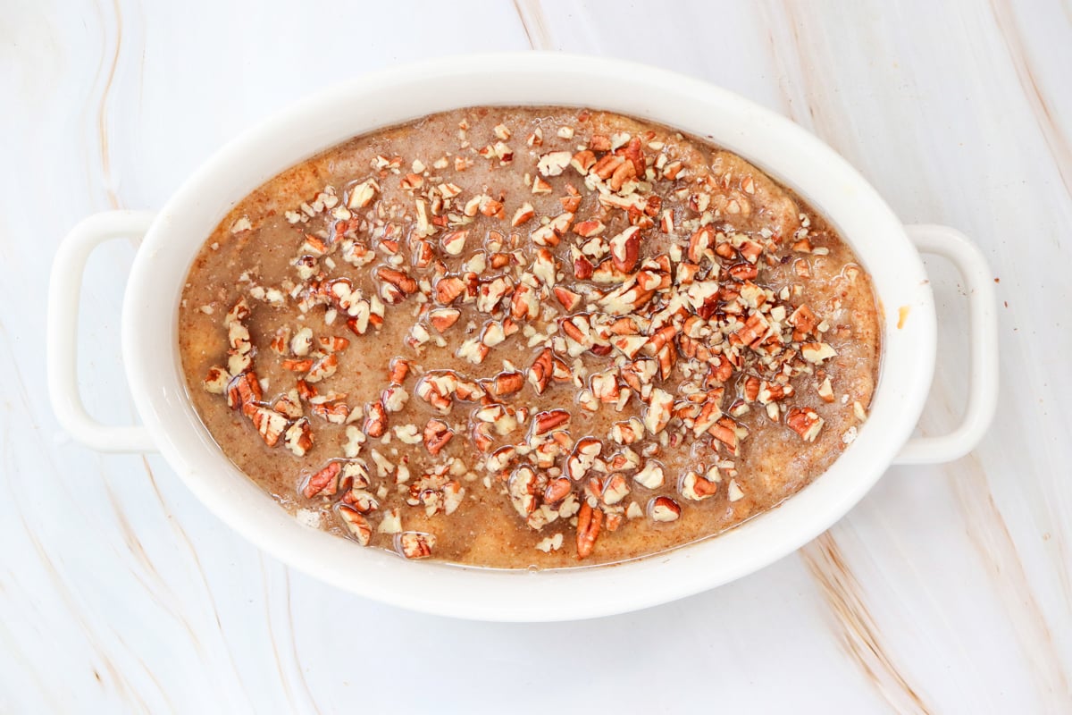 A white baking dish filled with a layer of dry ingredients for a gluten free pumpkin dump cake is placed on a marble countertop alongside a small bowl of chopped pecans and a can.
