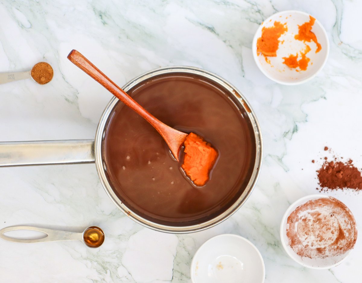 A pot of hot chocolate with a wooden spatula stirring in pumpkin puree, surrounded by small bowls of spices and ingredients for fall pumpkin hot chocolate recipes.
