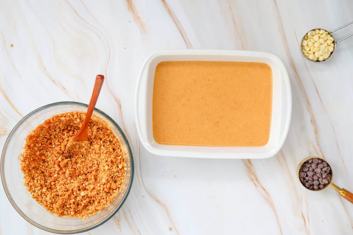 A white baking dish filled with an unbaked pumpkin crisp mixture sits on a marble surface, accompanied by measuring cups of white and dark chocolate chips.
