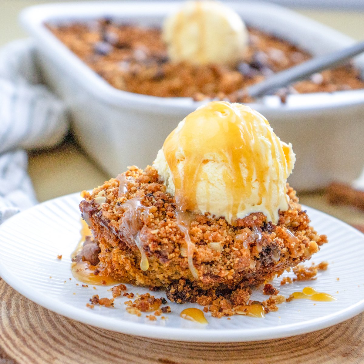 A serving of easy pumpkin crisp topped with vanilla ice cream and a drizzle of caramel sauce is placed on a white plate, with a baking dish of the pumpkin crisp in the background.