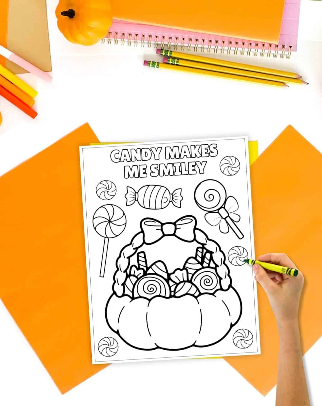 A hand holding a yellow crayon colors a printed Halloween coloring page featuring a pumpkin filled with candy, surrounded by orange stationery and decorative items.
