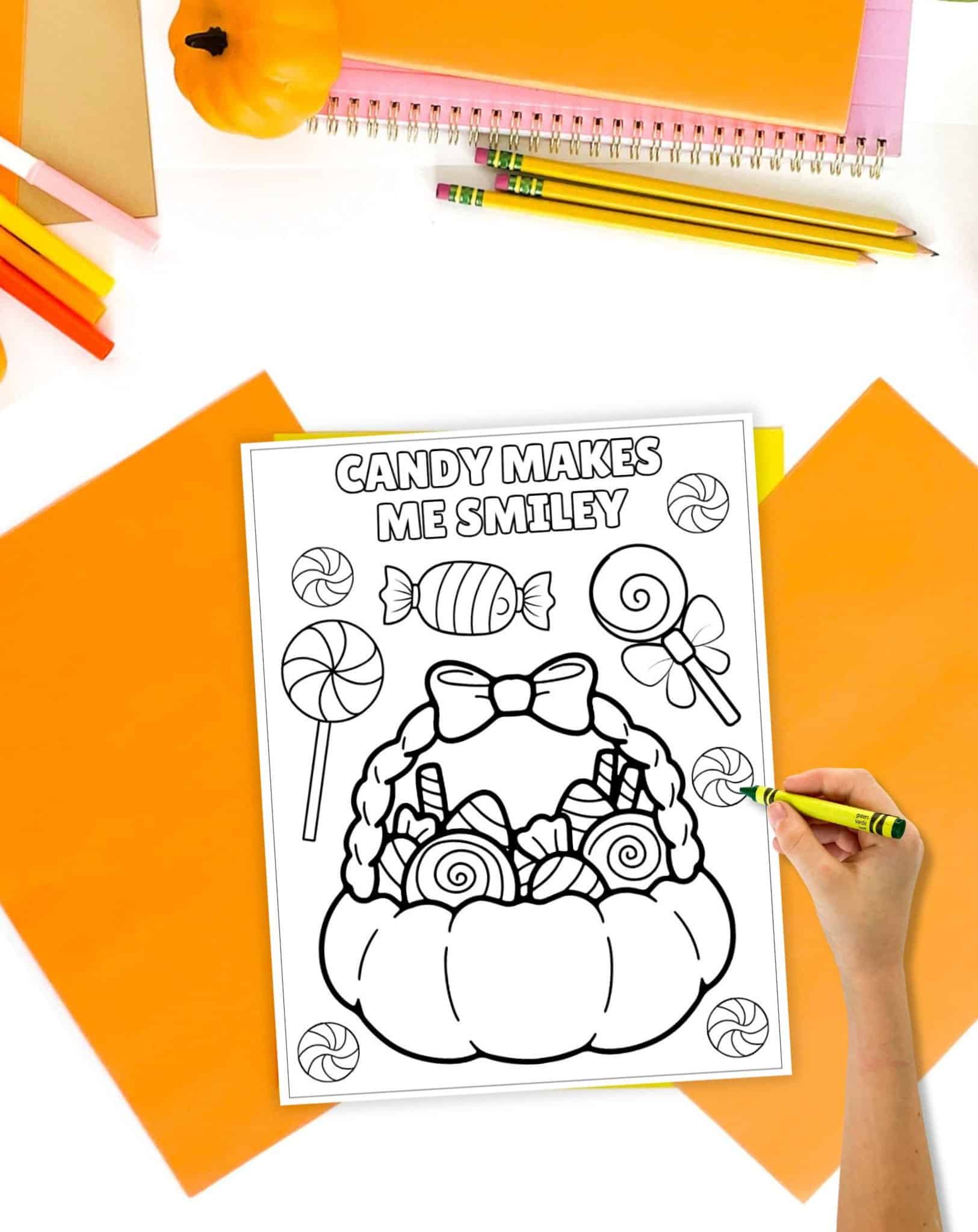 A hand holding a yellow crayon colors a printed Halloween coloring page featuring a pumpkin filled with candy, surrounded by orange stationery and decorative items.
