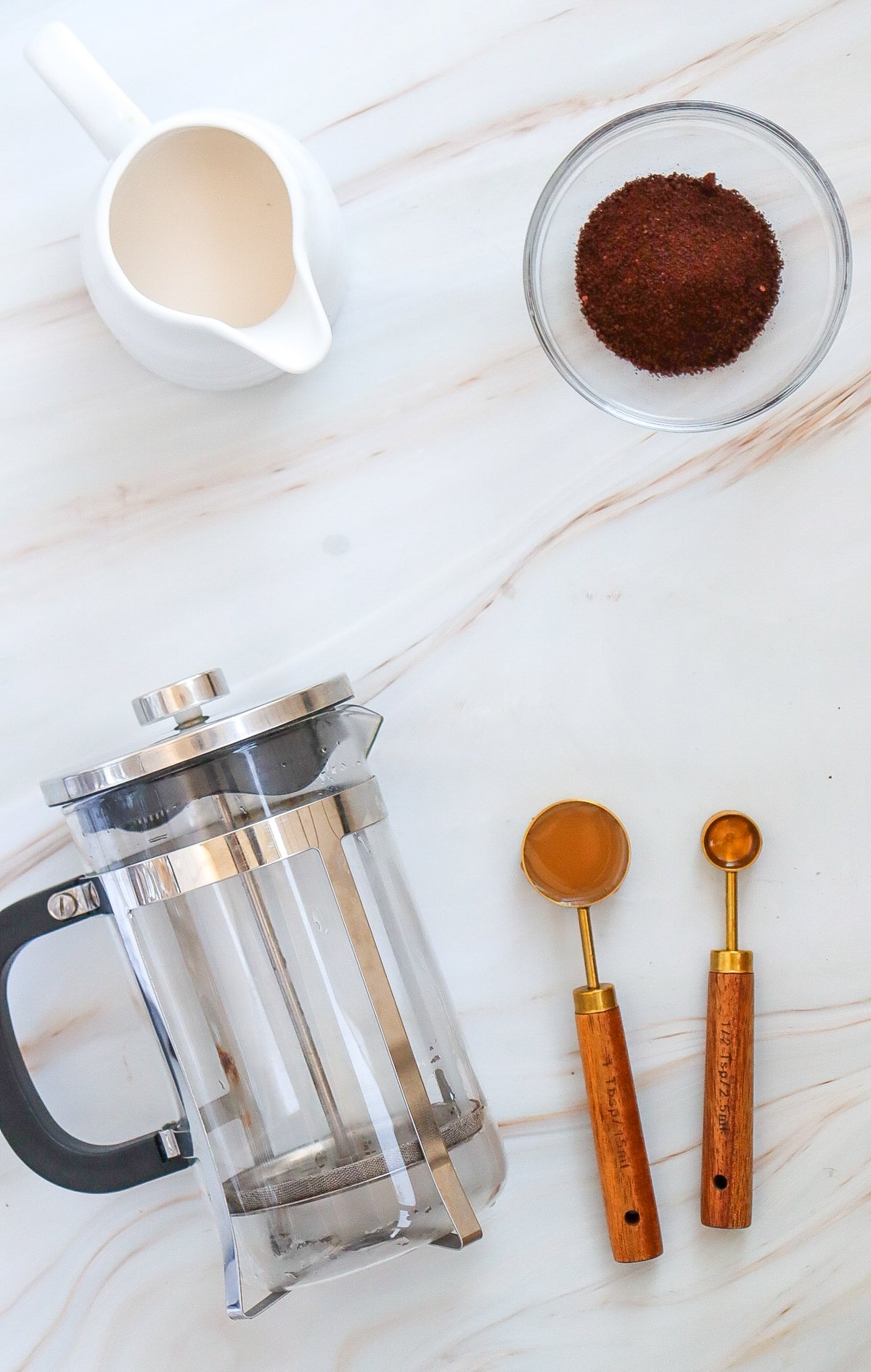 A French press, measuring spoons, a dish of coffee grounds, and a small milk jug are arranged on a marble surface, suggesting preparation for a homemade caramel iced coffee recipe.
