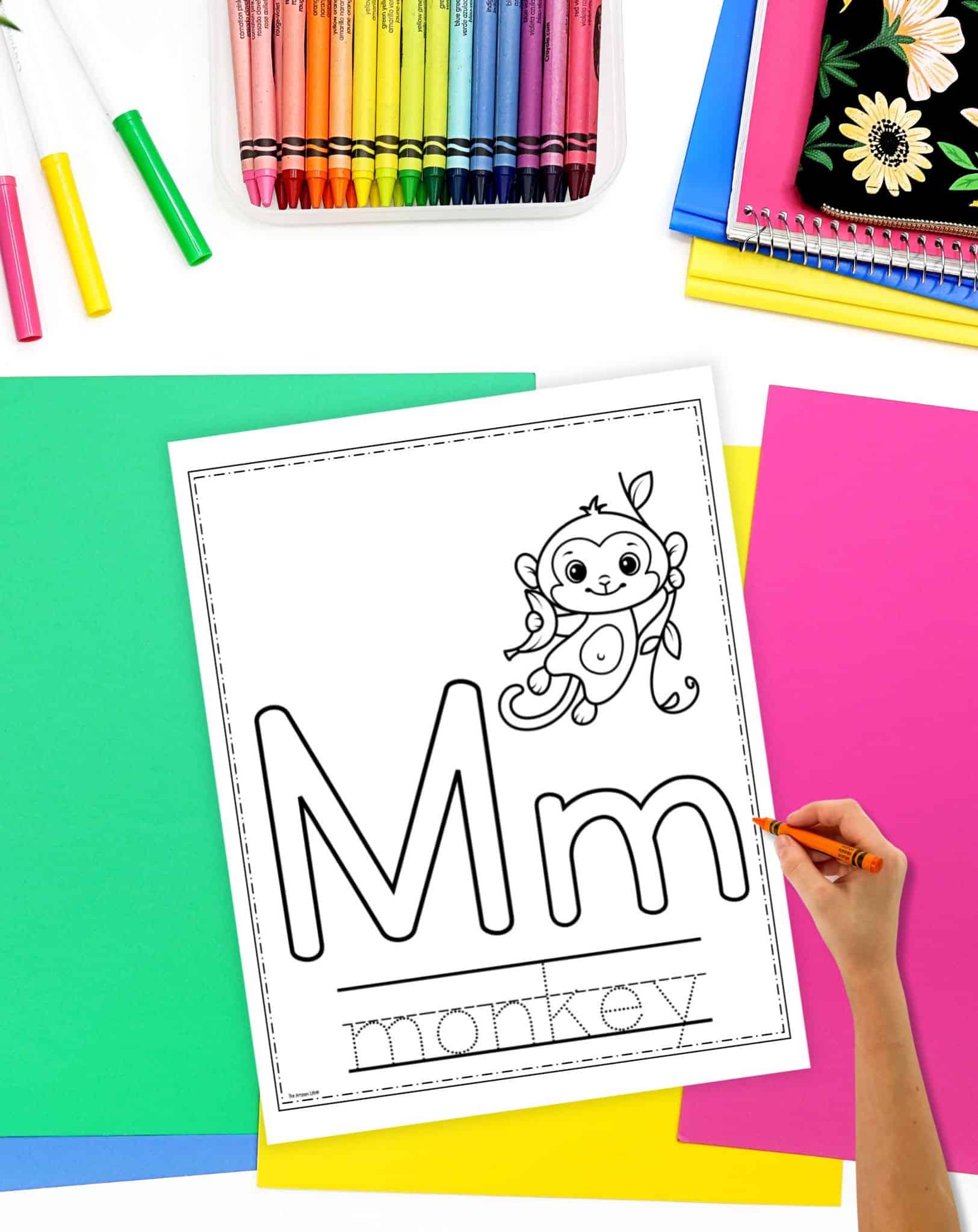 A hand is coloring a printable alphabet coloring activity page featuring the letter "M" and the word "monkey," surrounded by colorful art supplies and paper.
