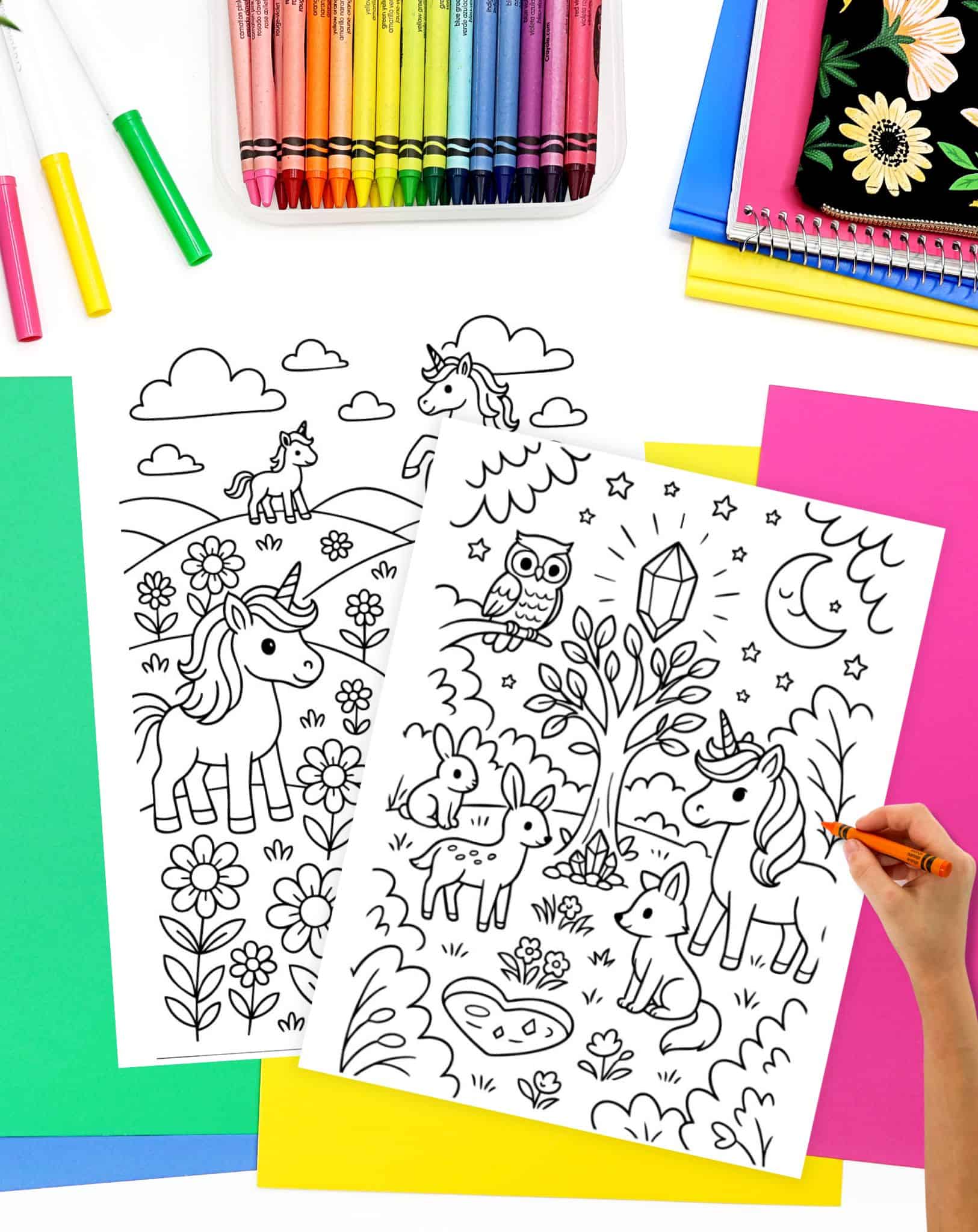 A hand is coloring a magical unicorn scene featured on a free printable unicorn coloring page, surrounded by additional coloring sheets, crayons, and colorful paper.
