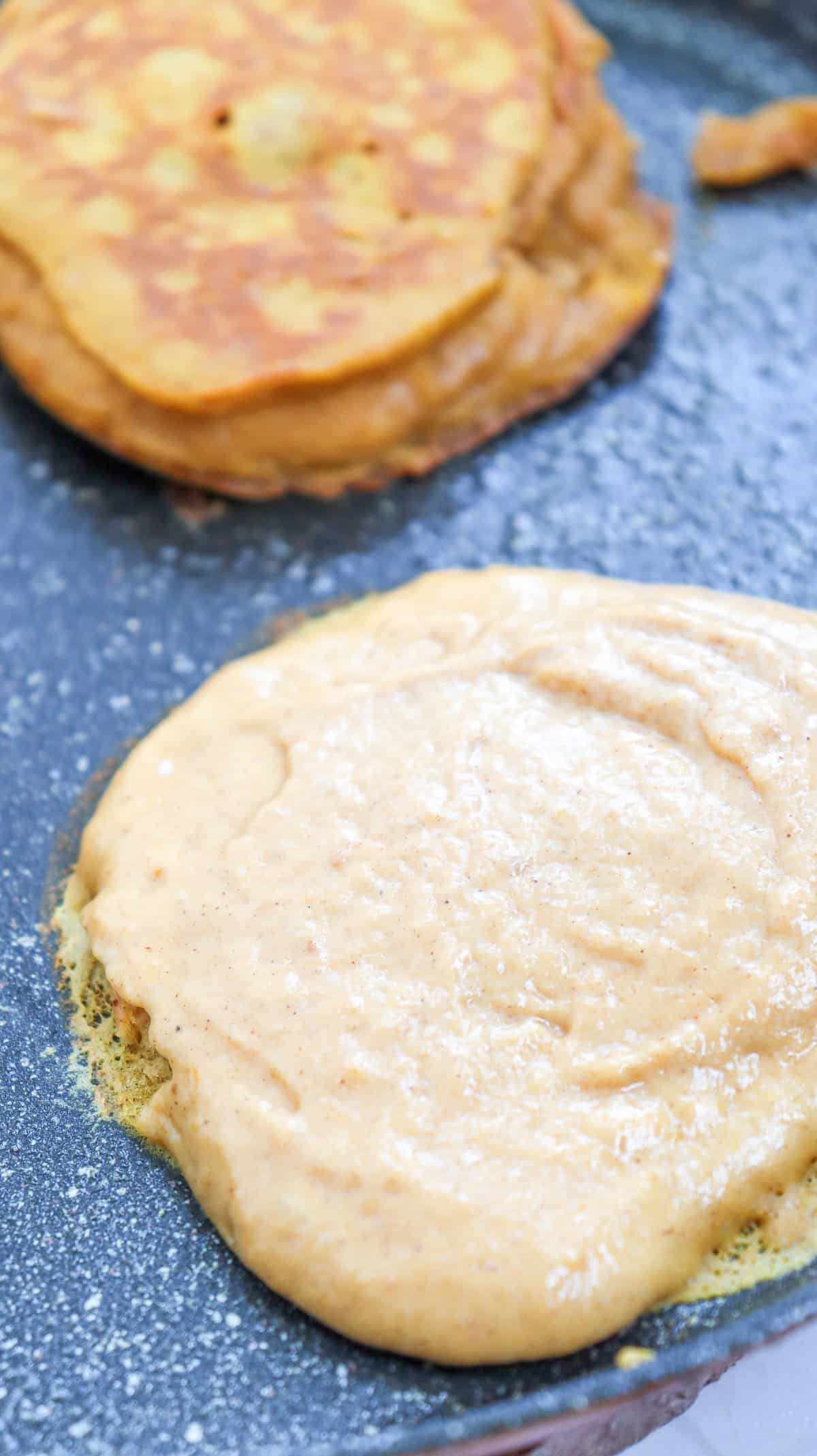 A close-up of a griddle with a blob of pumpkin pancake batter cooking alongside a fluffy cooked pumpkin pancake, highlighting a quick pumpkin pancake recipe.