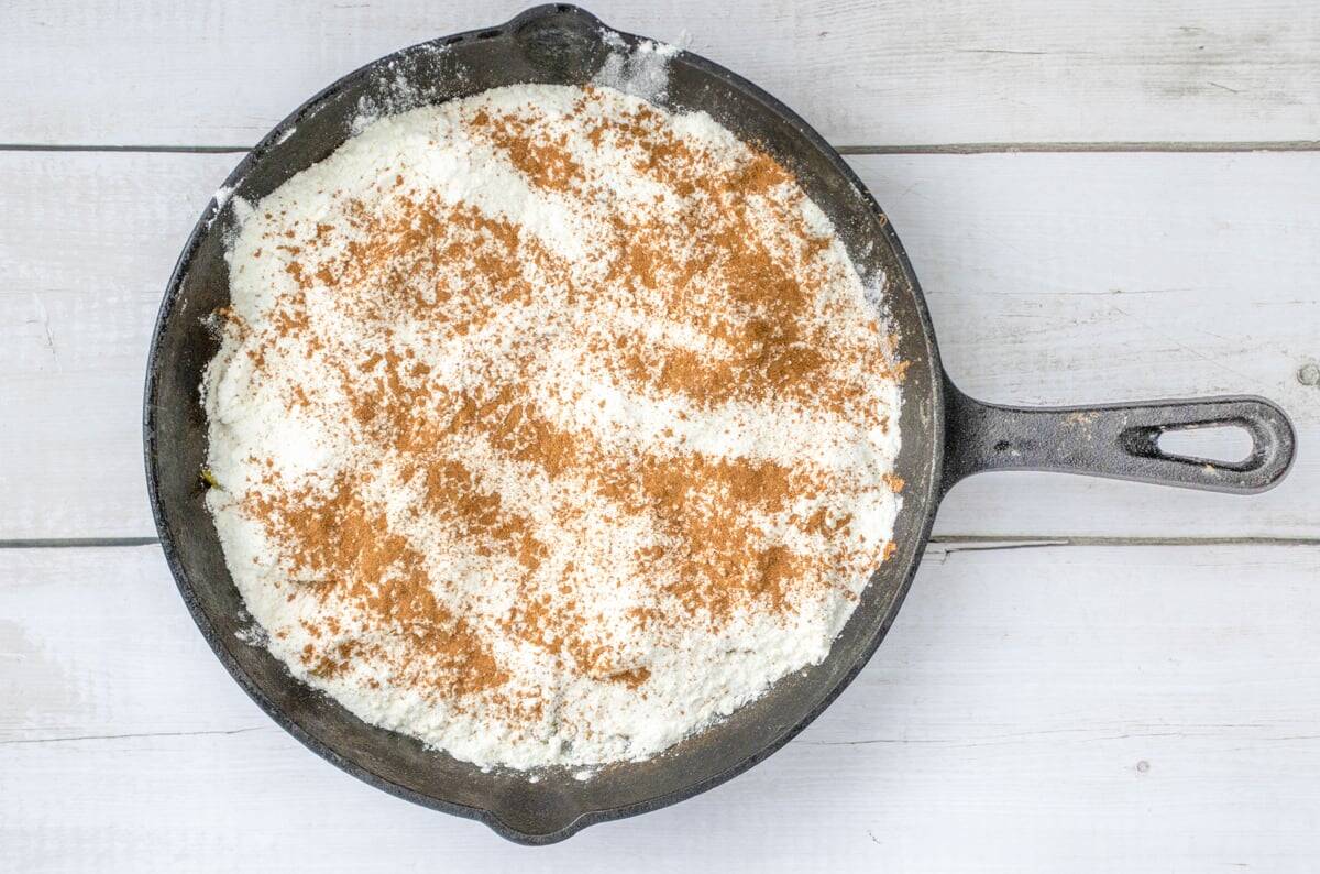 A cast-iron skillet contains a layer of ingredients for a fresh peach dump cake, topped with a sprinkling of cinnamon on a wooden surface.