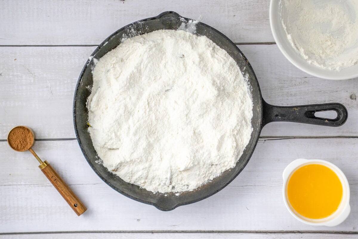 An overhead view of a cast iron skillet filled with cake mix, a teaspoon of cinnamon, and a small bowl of melted butter, set on a wooden surface, indicating preparation for a fresh peach dump cake.