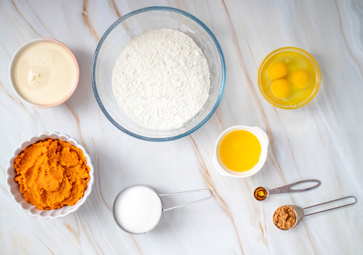Ingredients for an easy pumpkin dump cake recipe are displayed, including pumpkin puree, flour, eggs, sugar, and spices, laid out on a marble countertop.
