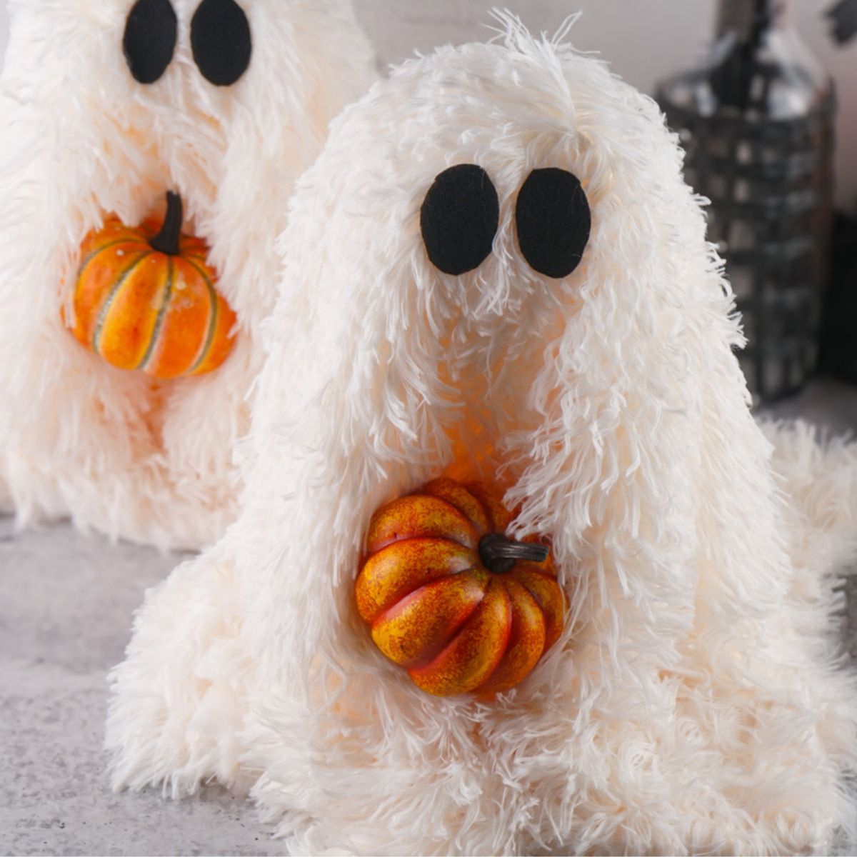 Two fluffy, white DIY ghosts, each holding a small orange pumpkin, are displayed as part of fall home decor, suitable for Halloween crafts.
