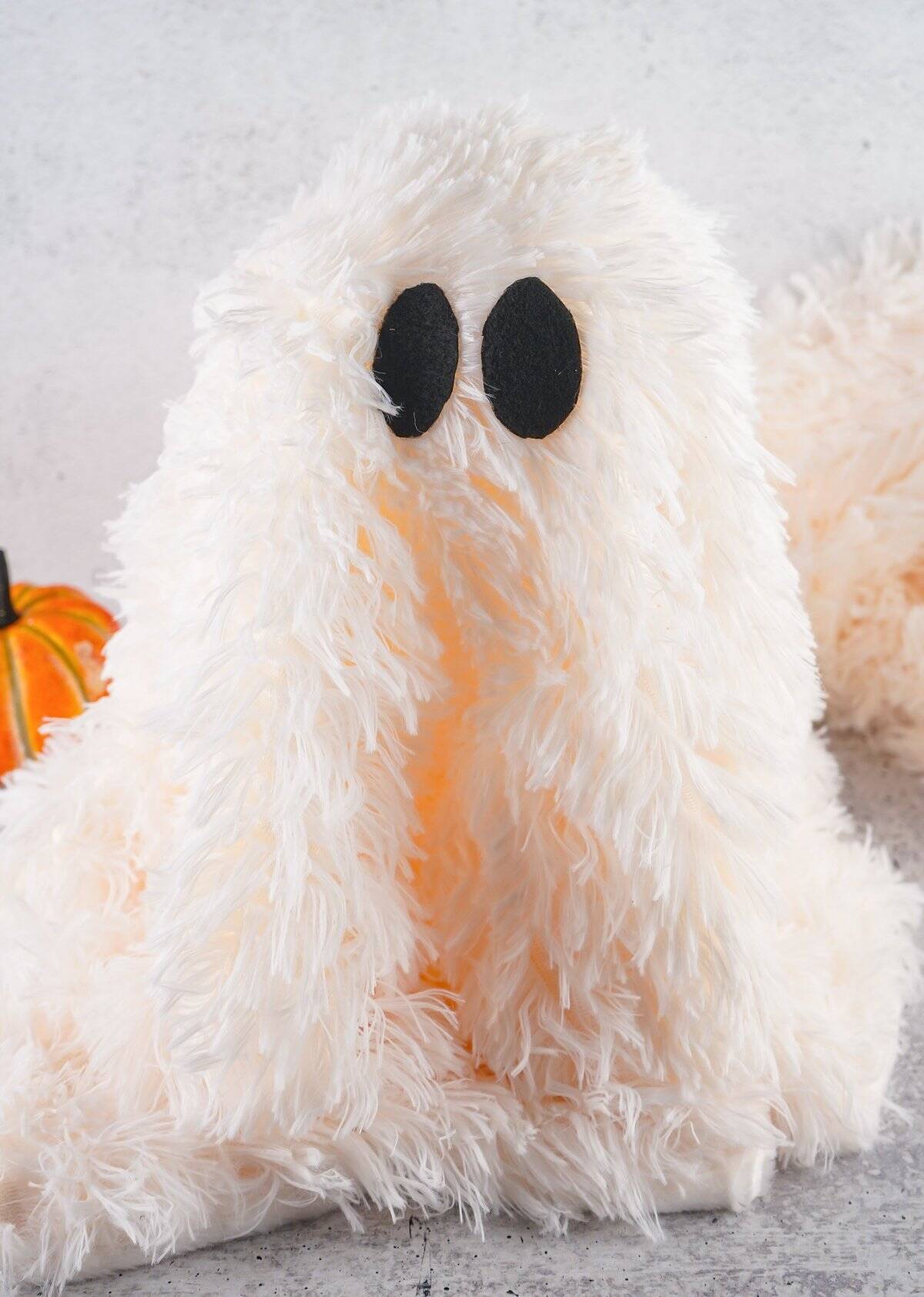 A hot glue gun is shown applying glue to two black felt pieces on a gray surface, with a fluffy white material and an orange decorative pumpkin in the background, suggesting a context of making fall and Halloween DIY decor such as a sherpa ghost.
