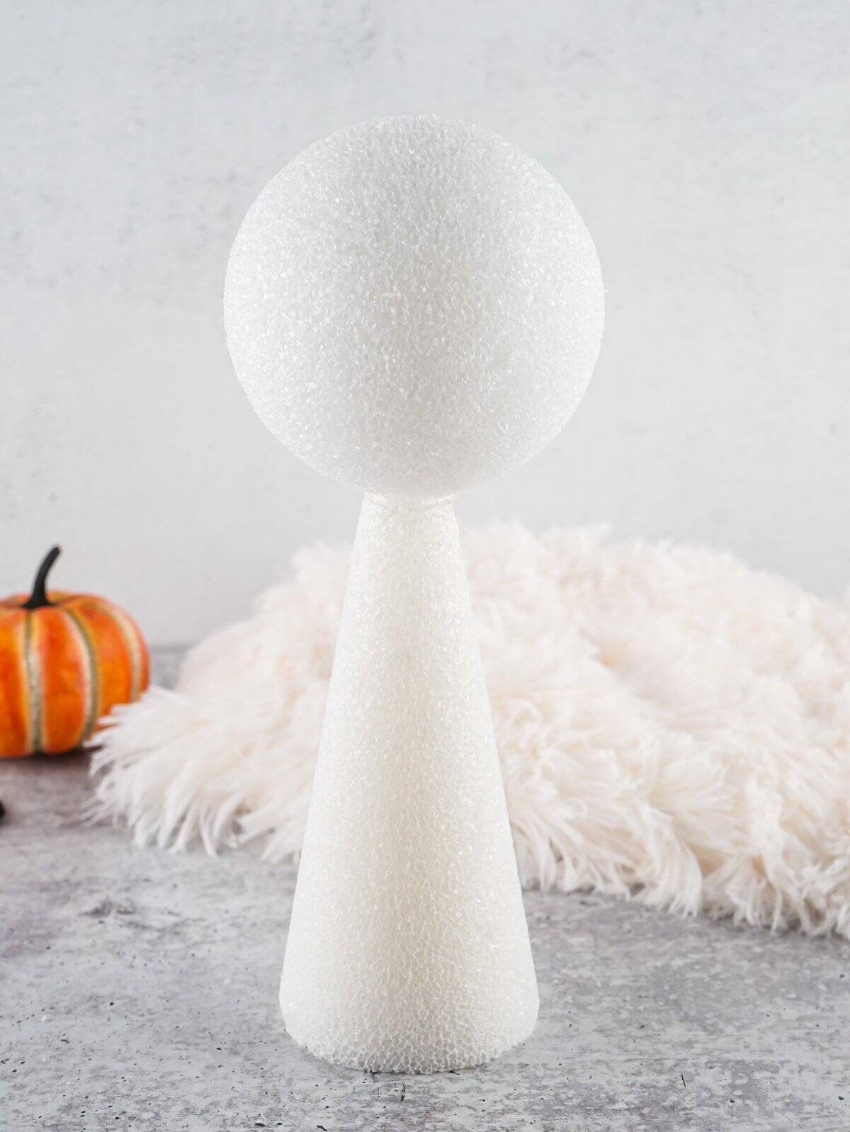 A styrofoam cone and ball are arranged to create a base for a DIY ghost decoration, set against a soft white backdrop and surrounded by seasonal decor elements.
