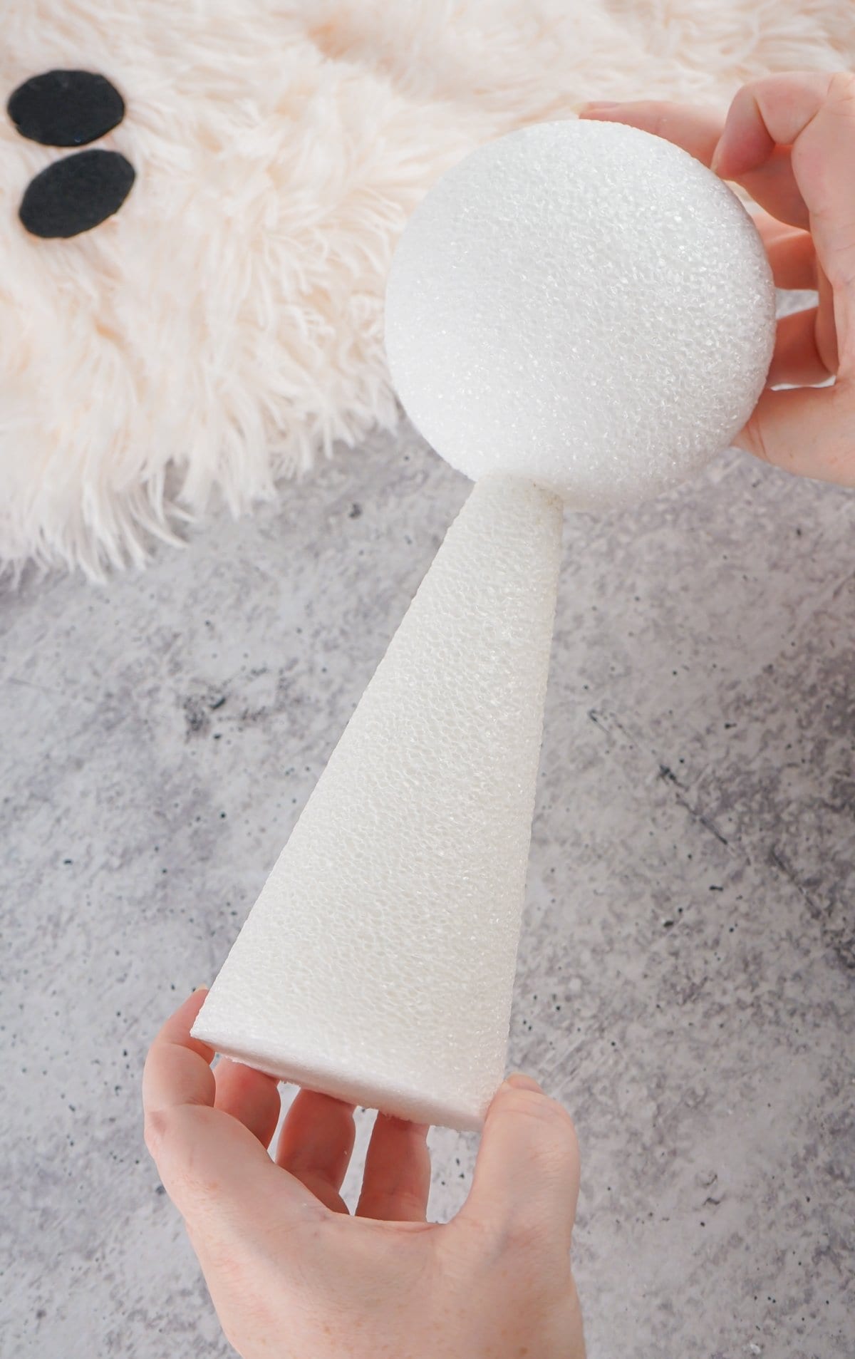 A person holds a white foam cone and sphere, intended for a ghost DIY project, on a gray surface with a fluffy white backdrop, illustrating a sherpa ghost decoration concept for fall and Halloween decor.


