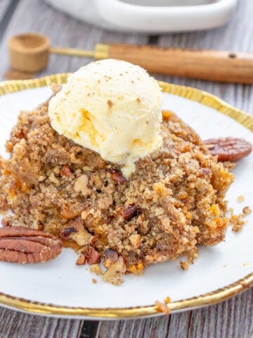A serving of gluten-free pumpkin dump cake topped with vanilla ice cream and garnished with pecans, with a baking dish of the same dessert in the background.