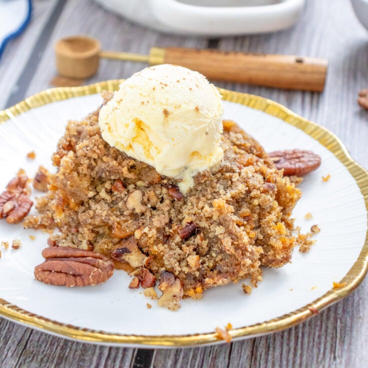 A serving of gluten-free pumpkin dump cake topped with vanilla ice cream and garnished with pecans, with a baking dish of the same dessert in the background.