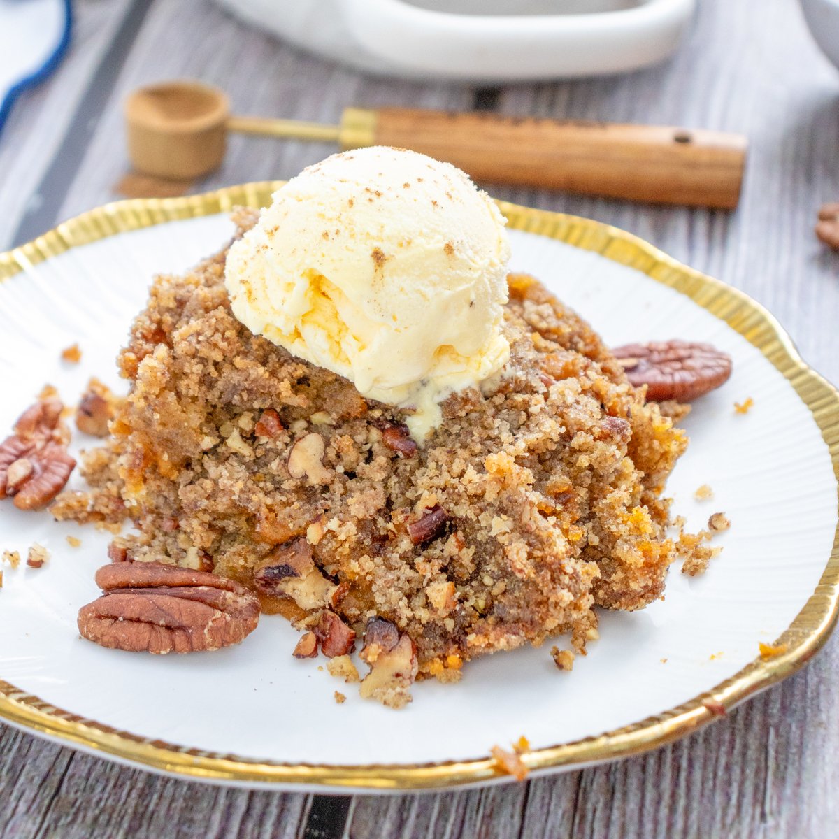A serving of gluten-free pumpkin dump cake topped with vanilla ice cream and garnished with pecans, with a baking dish of the same dessert in the background.
