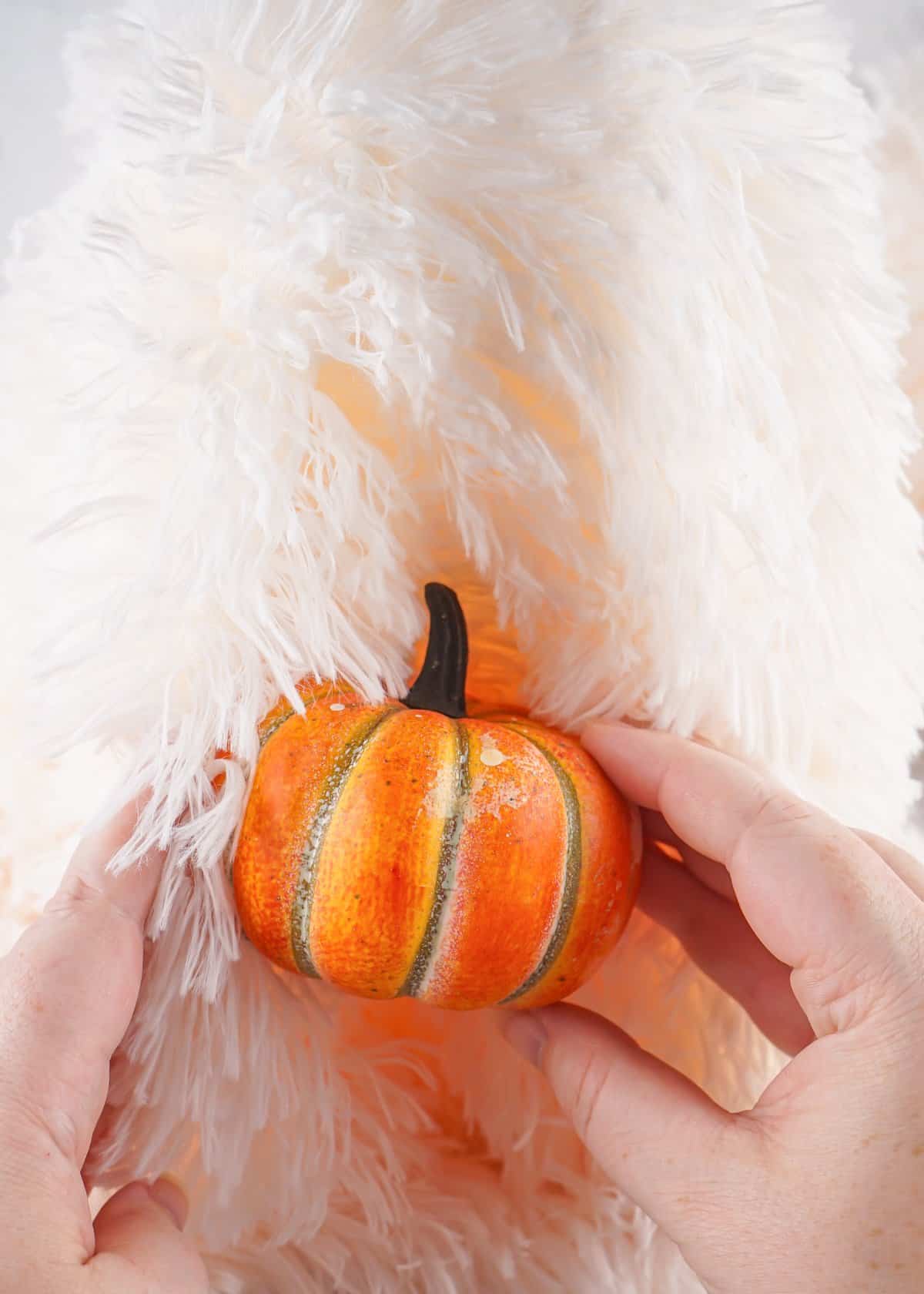 A person uses a hot glue gun to apply glue on an orange decorative pumpkin, with a soft, fluffy background suitable for fall decor DIY projects.
