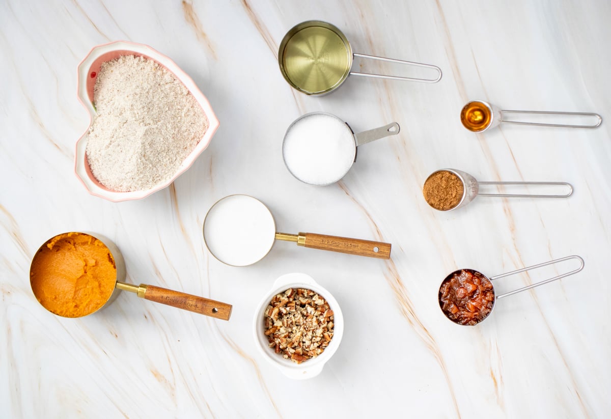 An assortment of measuring cups and ingredients for gluten free pumpkin dump cake, including flour, sugar, pumpkin puree, and chopped nuts, arranged on a marble surface.
