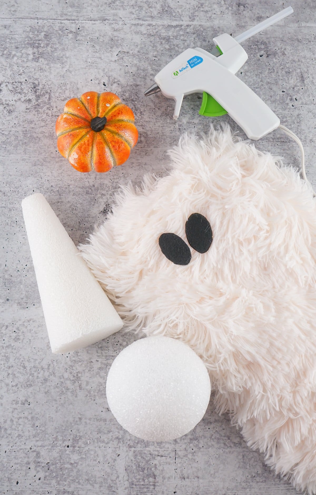 A flat lay image featuring a fluffy white fabric, black felt eyes, two Styrofoam forms (a cone and a sphere), a small orange pumpkin, and a hot glue gun, suggesting materials for a DIY sherpa ghost craft for fall home decor.

