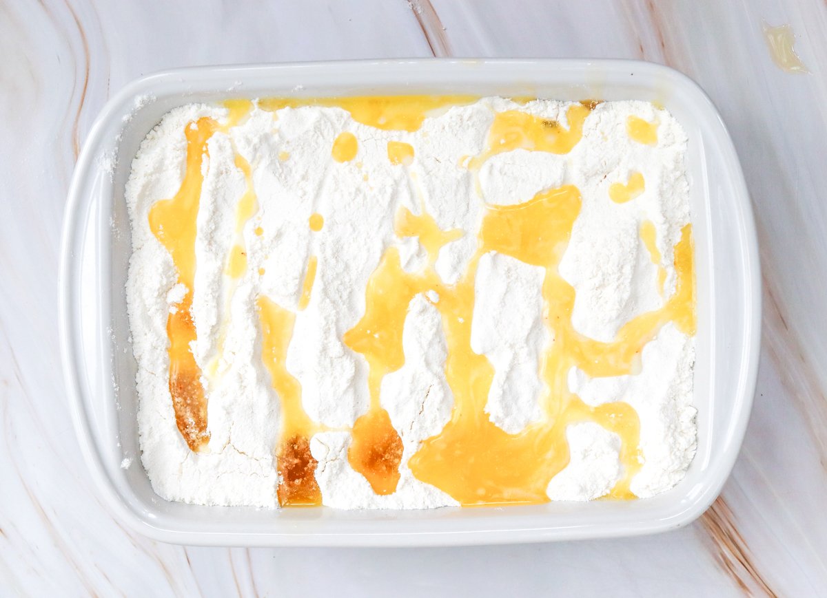A baking dish containing a layer of flour topped with drizzles of melted butter, illustrating the preparation of an easy pumpkin dump cake recipe.
