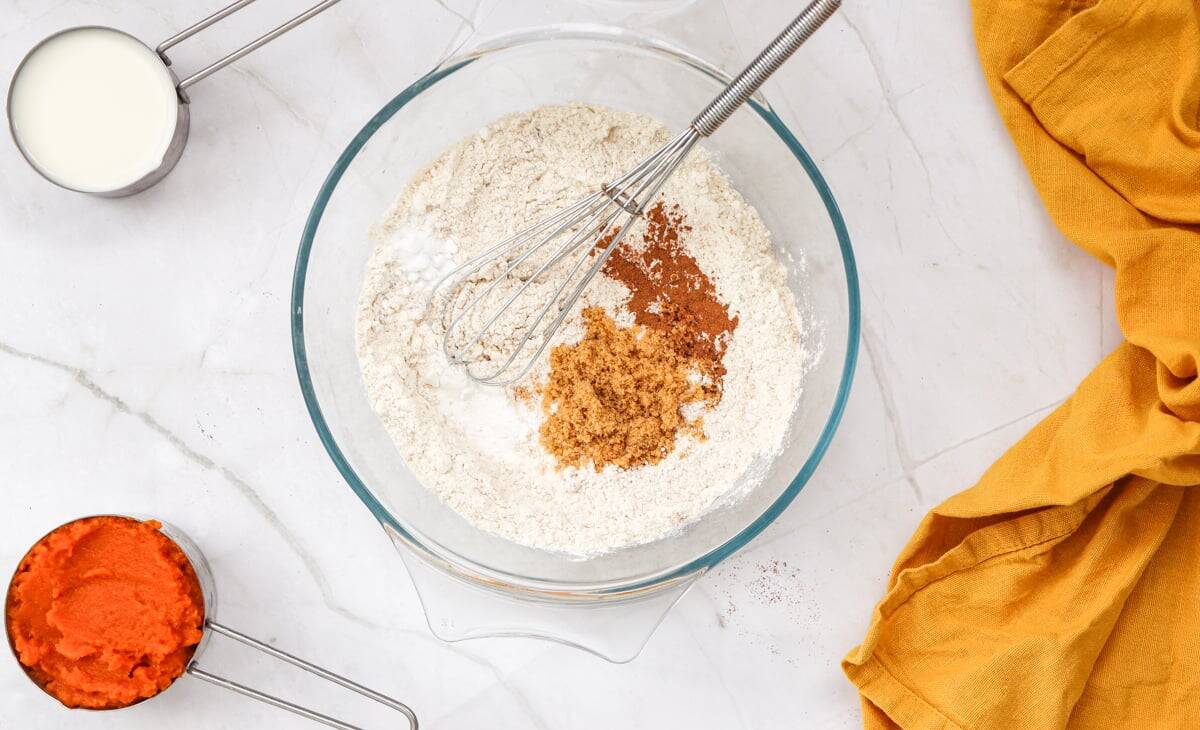 A glass bowl contains a mixture of flour, brown sugar, and cinnamon, with a whisk resting inside, accompanied by a measuring cup of milk and a bowl of pumpkin puree, set against a marble surface and a yellow cloth.