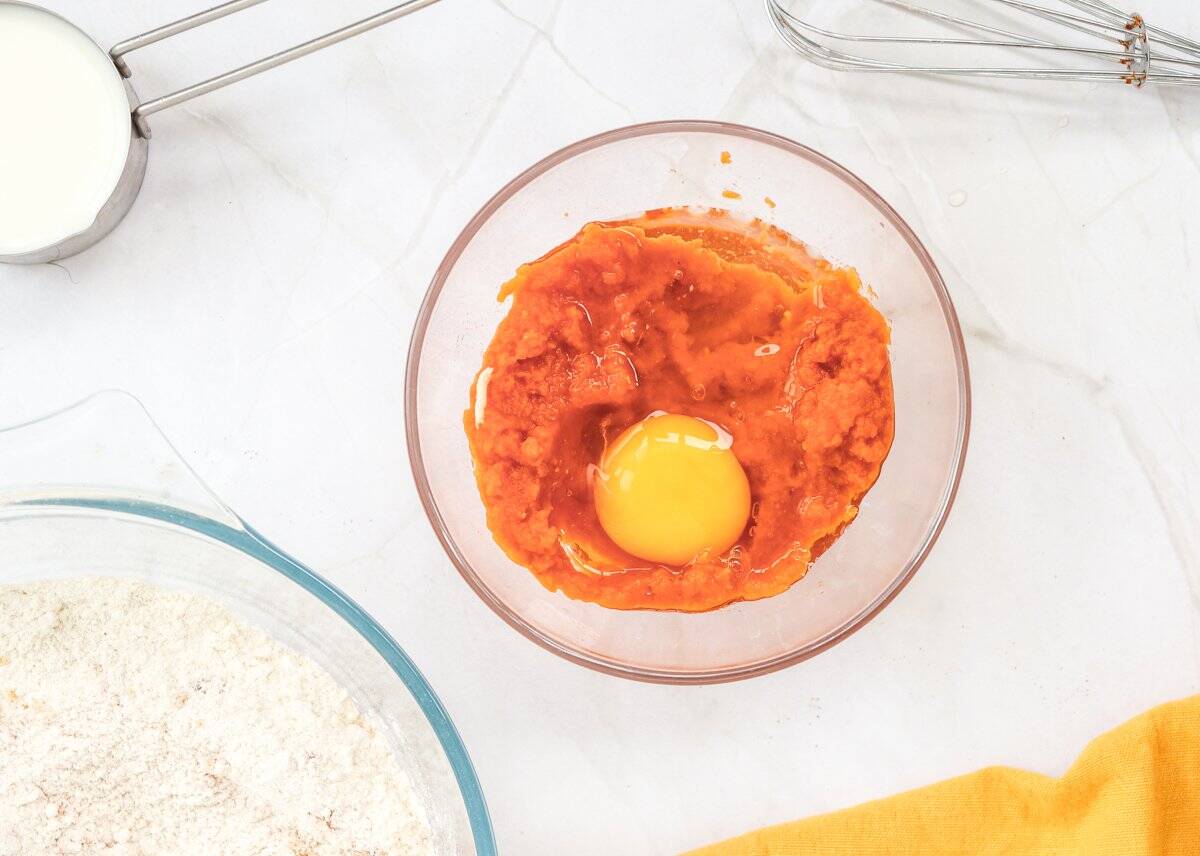 A bowl containing pumpkin puree and an egg is surrounded by ingredients for a quick pumpkin pancake recipe, including flour and milk, on a white countertop.